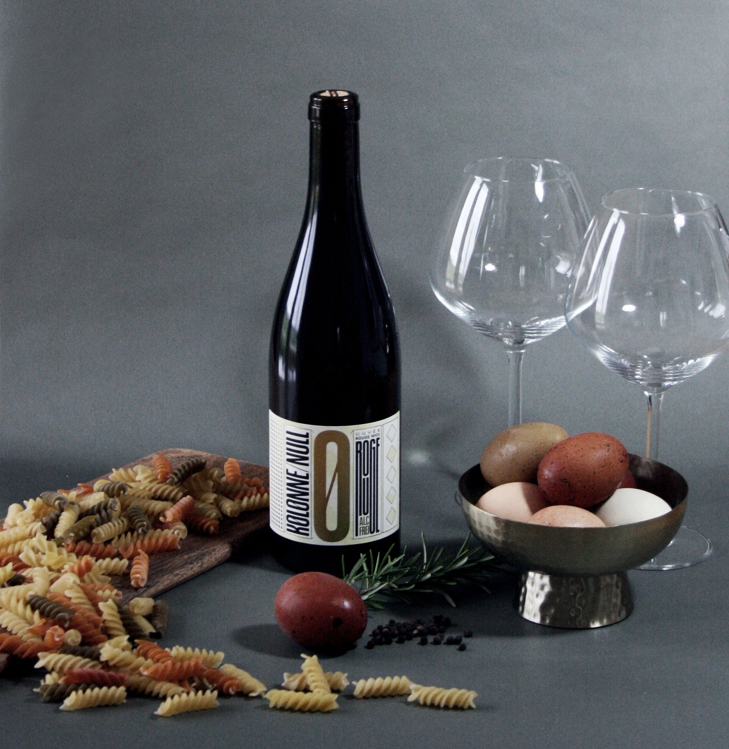 Wine bottle with a label, two wine glasses, and a bowl of fruits on a gray surface.