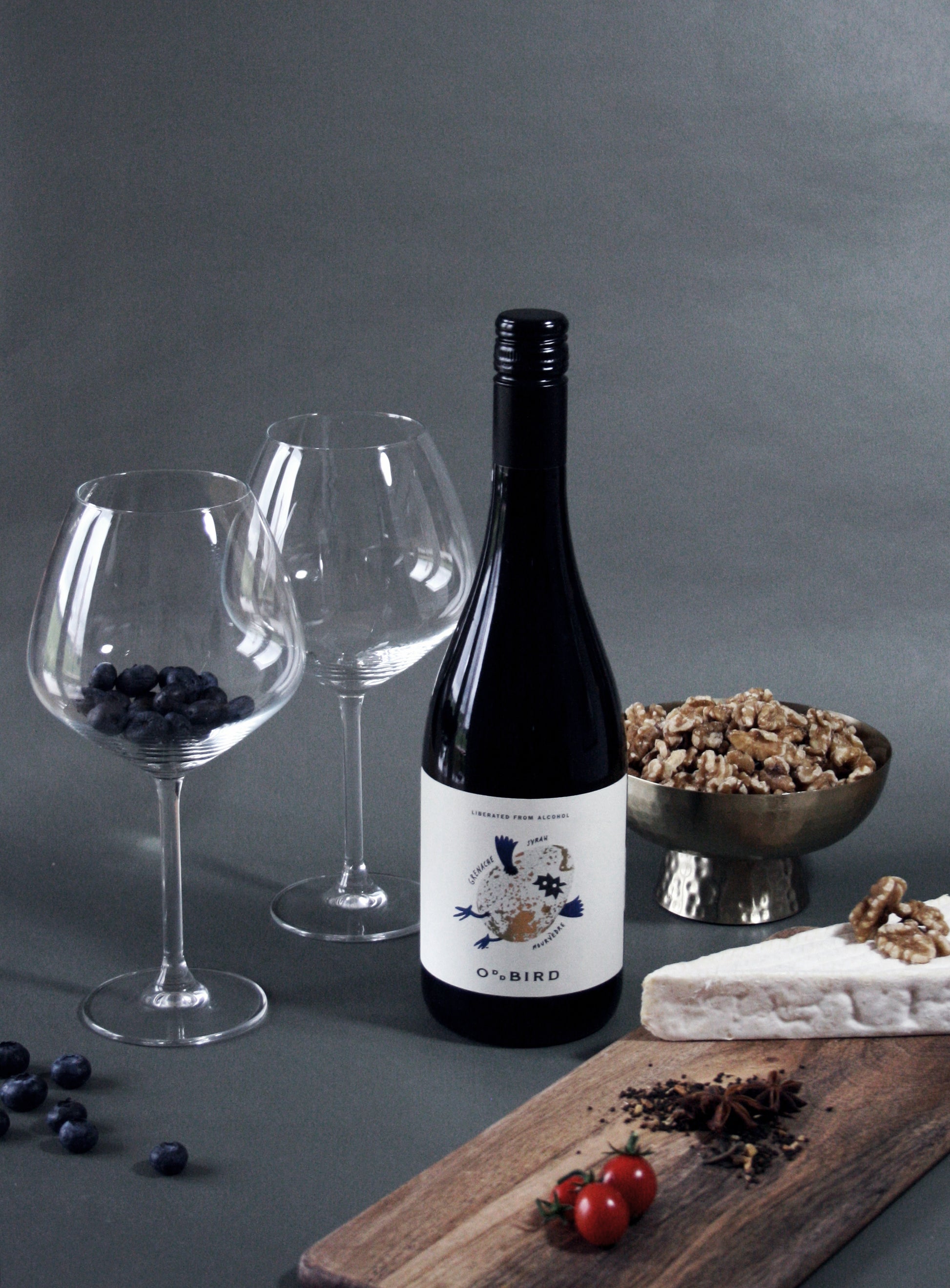 Wine bottle with two glasses, a bowl of nuts, and a cutting board with cheese and fruits on a gray background