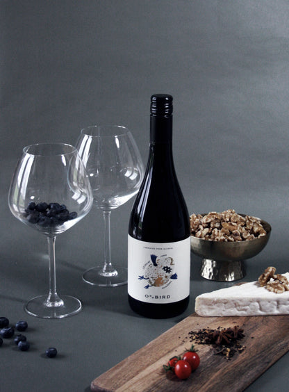 Wine bottle with two glasses, a bowl of nuts, and a cutting board with cheese and fruits on a gray background