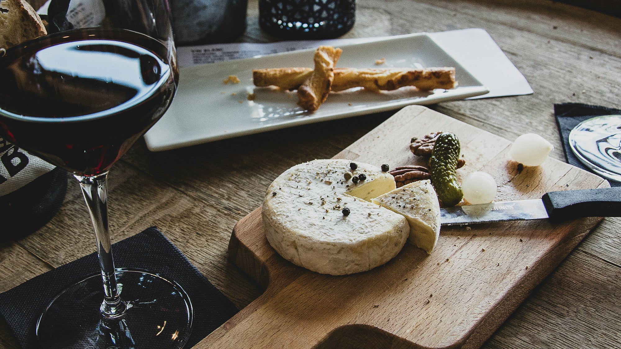 Wooden cutting board with cheese and crackers next to a glass of red wine on a wooden table. Photo by Camille Brodard on Unsplash